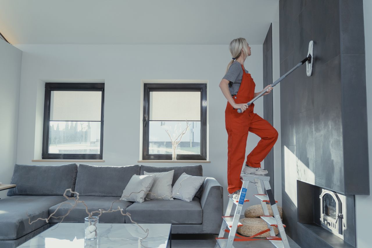 A woman in overalls cleans a wall in a stylish living room, enhancing home hygiene.